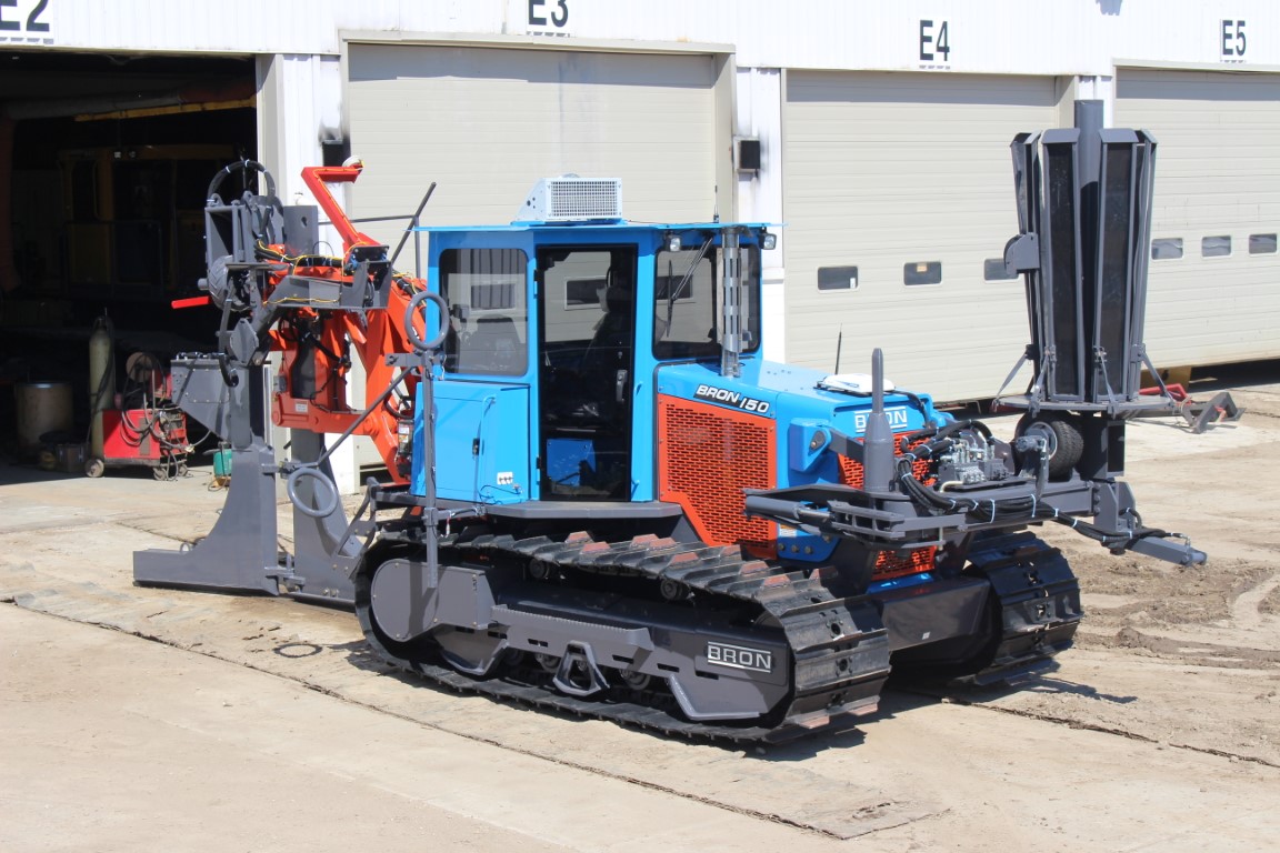 BRON V100 utility tractor equipped with various attachments, including a blue and orange plow unit, parked outside a service garage with loading docks in the background.