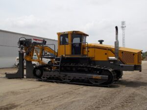BRON 350 utility tractor with heavy-duty plow and cable laying equipment, parked on a gravel lot next to an industrial building.