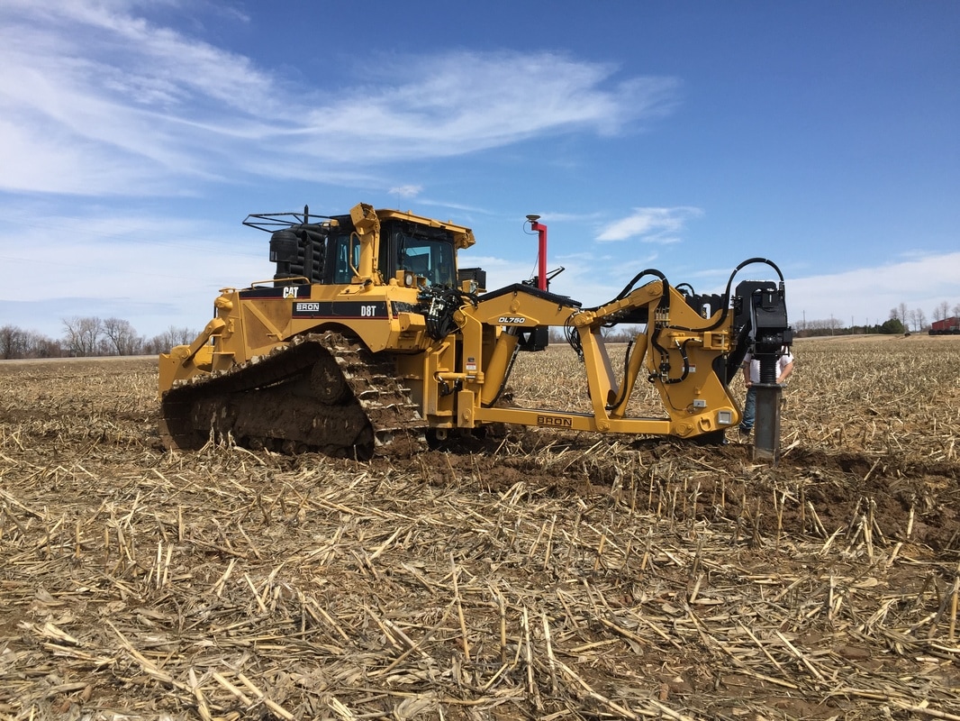 BRON utility tractor with attached plowing and trenching equipment, operating in a harvested field under a clear blue sky.